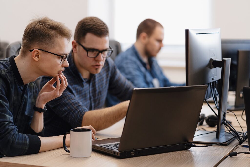 Two male colleagues are sitting at a desk and looking intently at a laptop screen. The man on the left is holding his chin in thought. A third colleague is out of focus in the background.