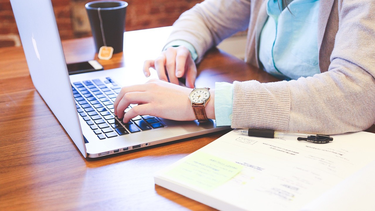 A person is typing on a silver laptop. A cup of tea and a stack of papers with sticky notes are visible on the wooden desk.