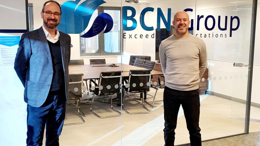 Two men are standing side by side in an office in front of a glass wall with the "BCN Group" logo and a conference room visible behind them. They are both smiling and have their hands clasped behind their backs.