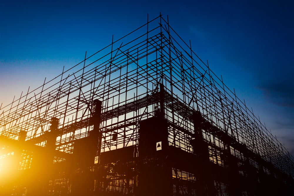 A silhouetted construction site at sunset, with a large scaffolding structure filling the frame against a deep blue and orange sky.