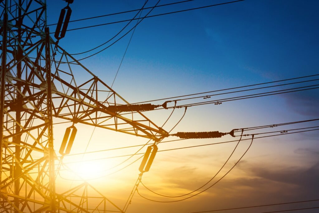The bottom of a power pylon with high-voltage wires against a sky transitioning from orange to blue at dusk.