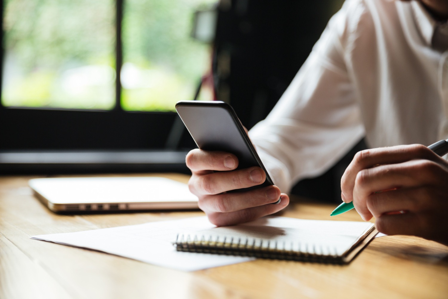A close-up shot of a person's hands holding a smartphone in one hand and a pen in the other, with a notebook on the table. A laptop is visible in the blurred background.