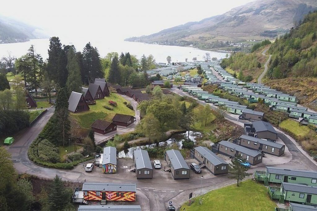 An aerial view of a caravan park and wooden lodges beside a loch surrounded by hills in a rural landscape.