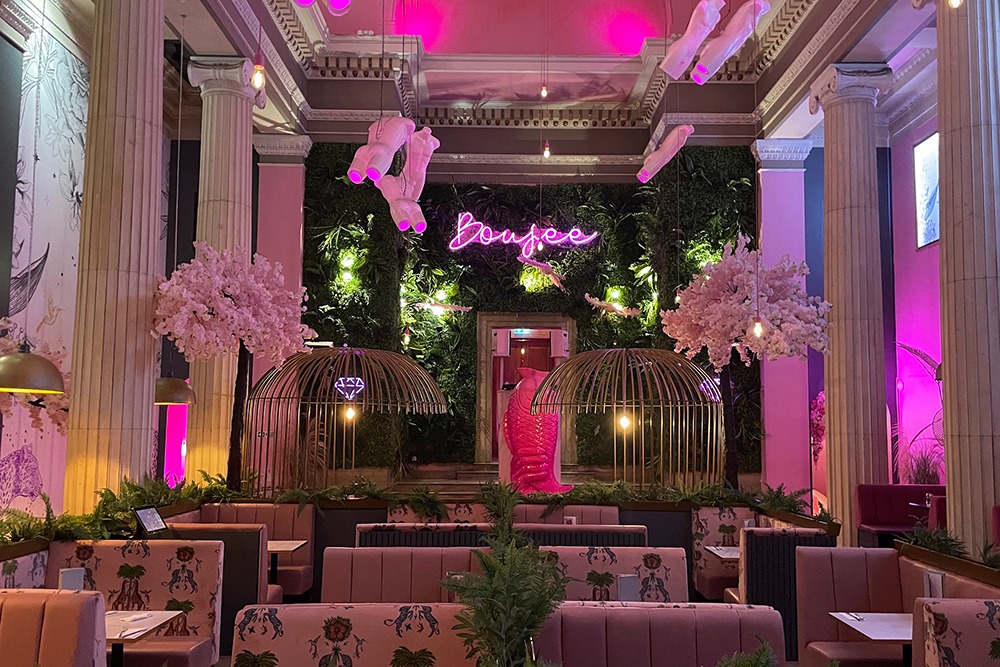 Boujee, an restaurant with a whimsical, pink-themed décor. The space features pink neon lighting, lush greenery on the walls, pink velvet booths, large birdcage decorations, and cherry blossom trees.