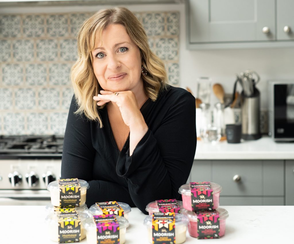 Julie Waddell, founder of Moorish, smiling and resting her chin on her hand, standing behind a kitchen counter with several containers of "Moorish Hummus" in front of her.
