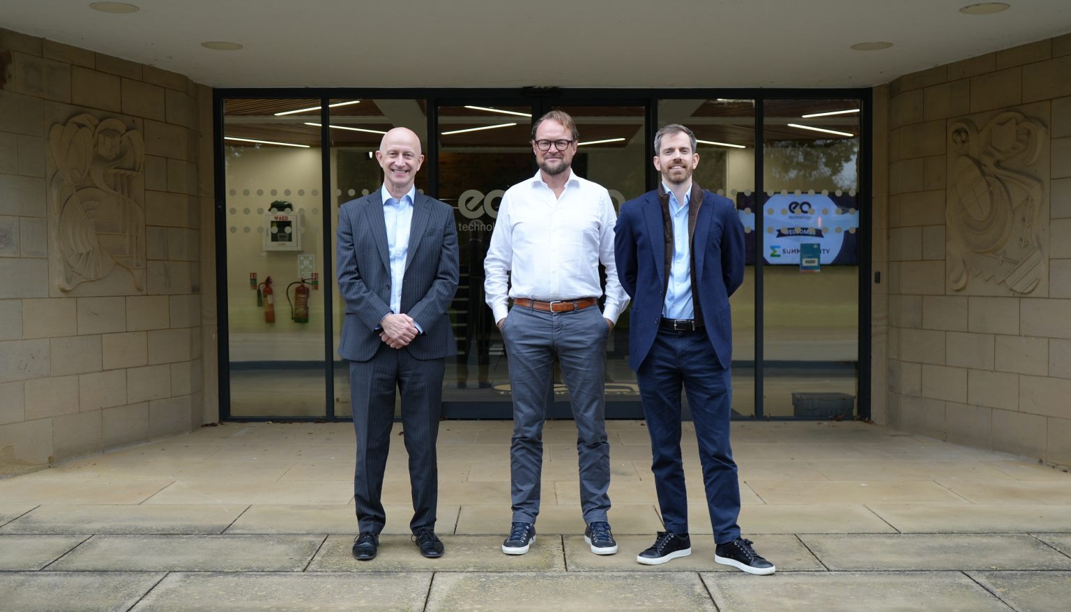 Three men are standing side by side in front of an office building with the "ECI Technology" logo on the door. All three are smiling and looking at the camera.