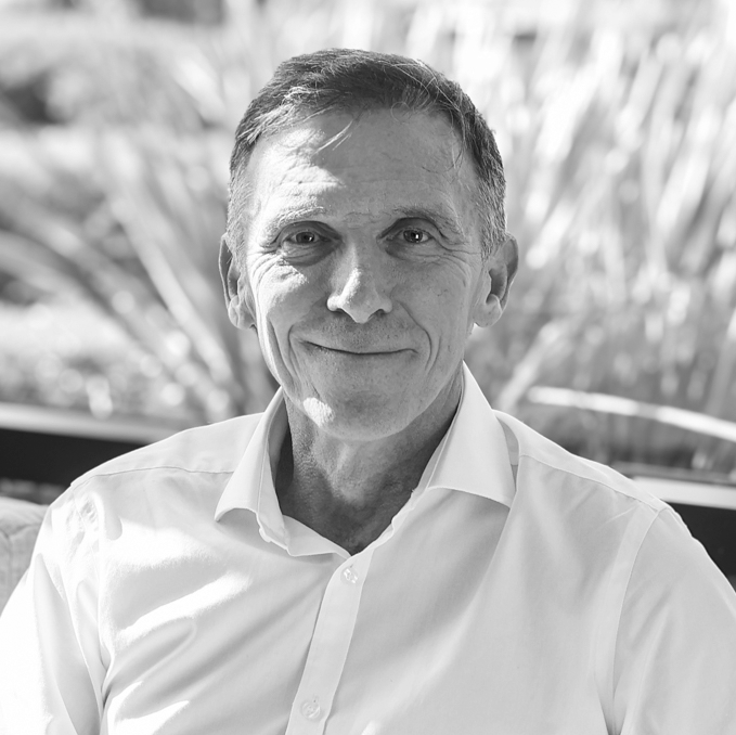 A black and white headshot image of a smiling man on a sofa in front of a window backdrop.