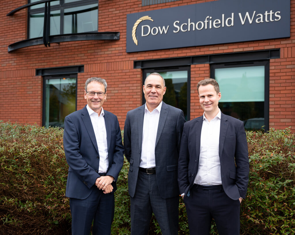 A group photo of three men smiling in front of the Dow Schofield Watts sign and building.
