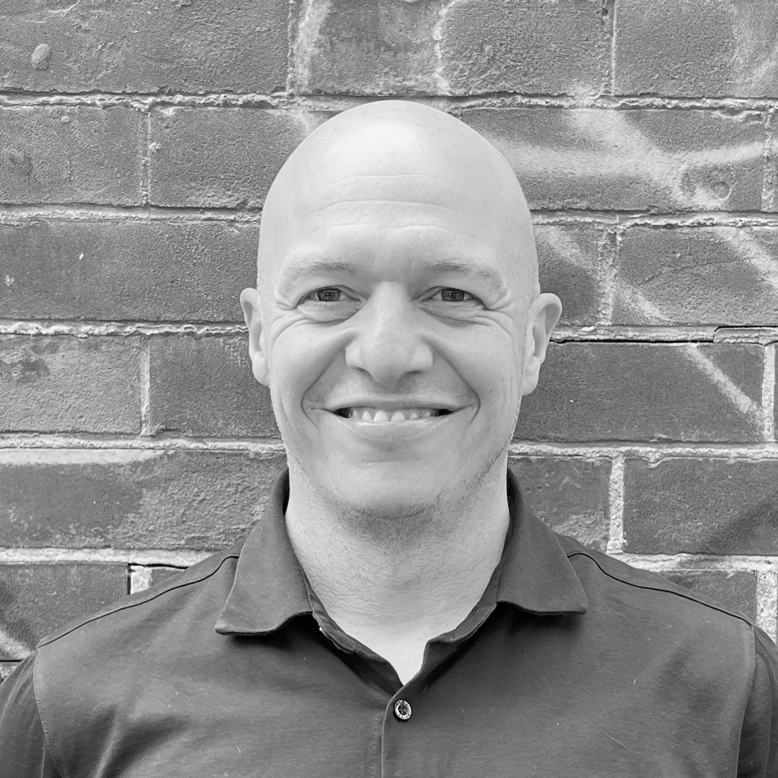 A black and white headshot image of a smiling man against a brick backdrop