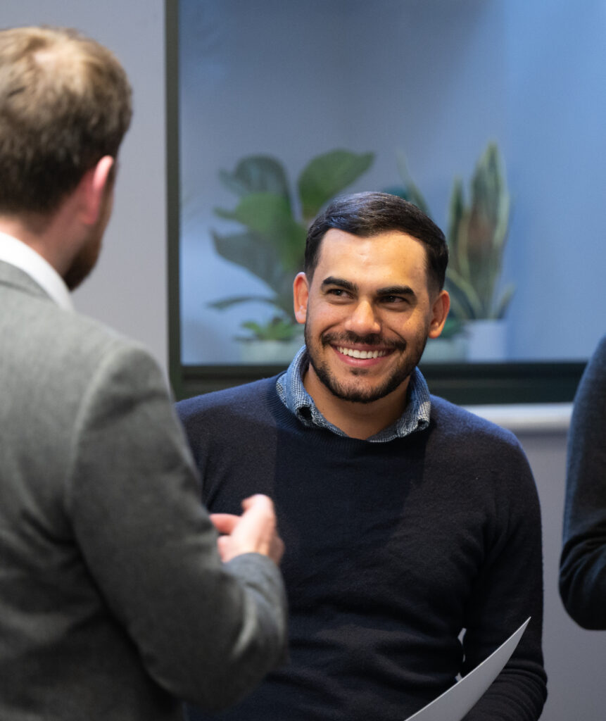 An image of a man smiling in conversation with another male, who has his back to the camera