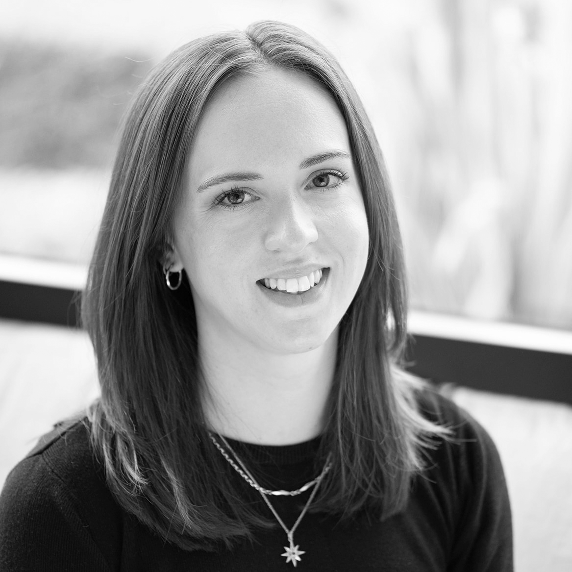 A black and white headshot image of a smiling woman on a sofa in front of a window backdrop.