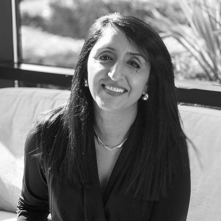 A black and white headshot image of a smiling woman on a sofa in front of a window backdrop.
