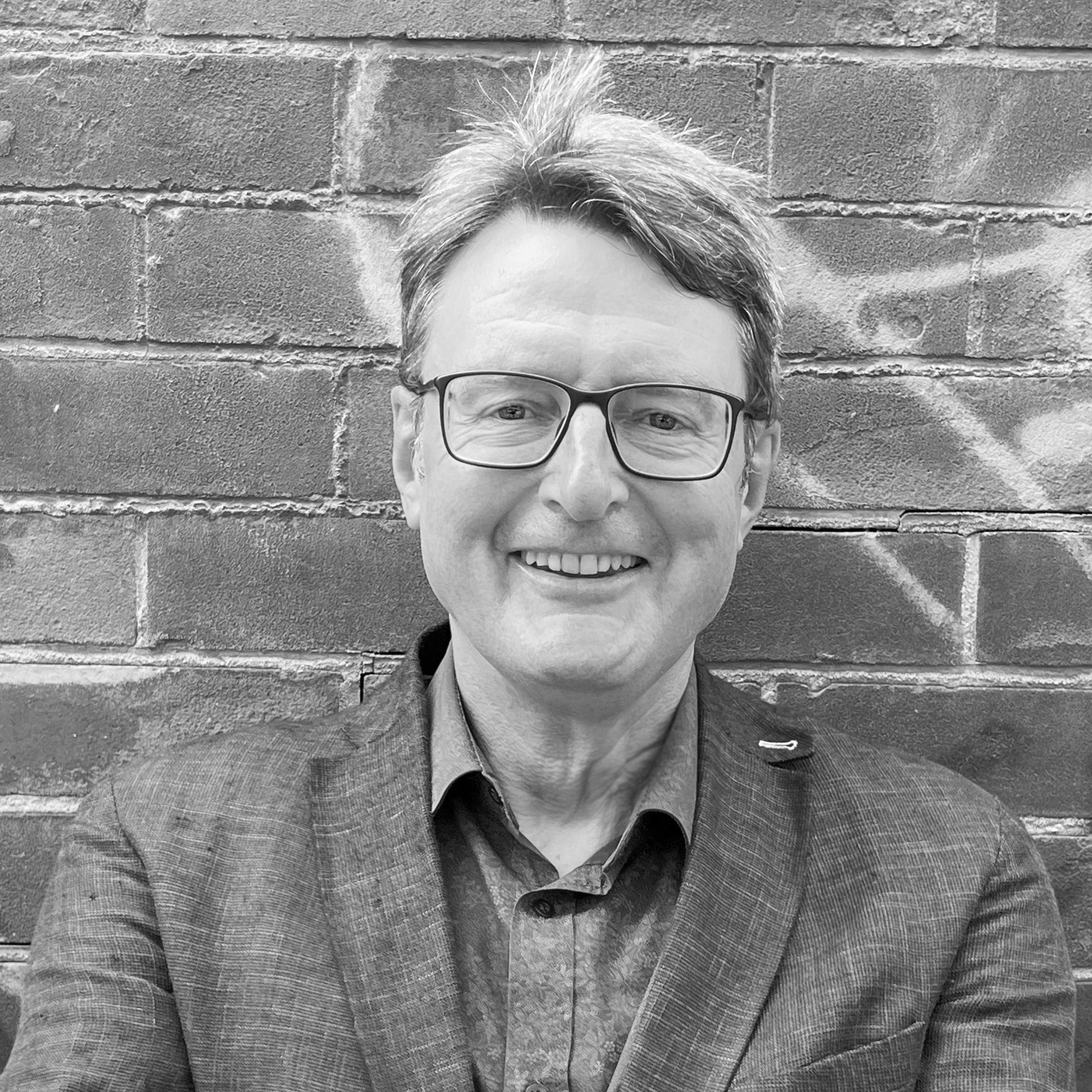 A black and white headshot image of a smiling man with glasses against a brick backdrop