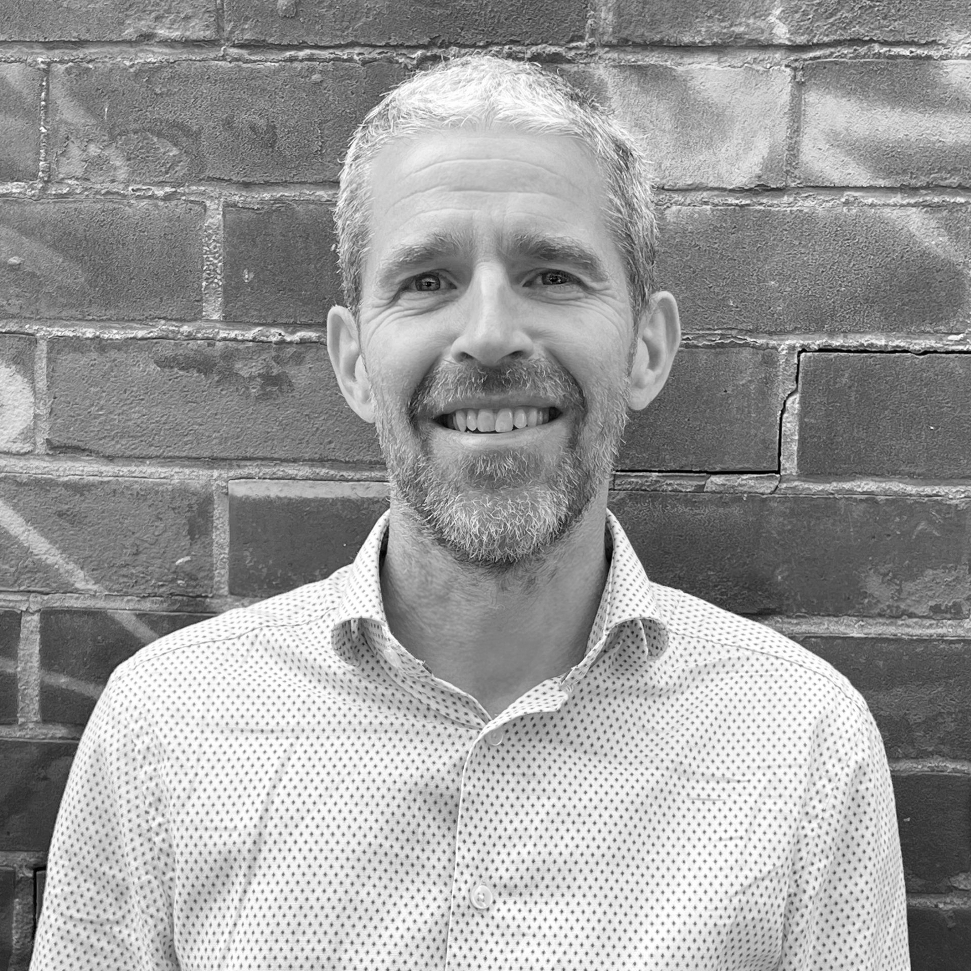 A black and white headshot image of a smiling man against a brick backdrop