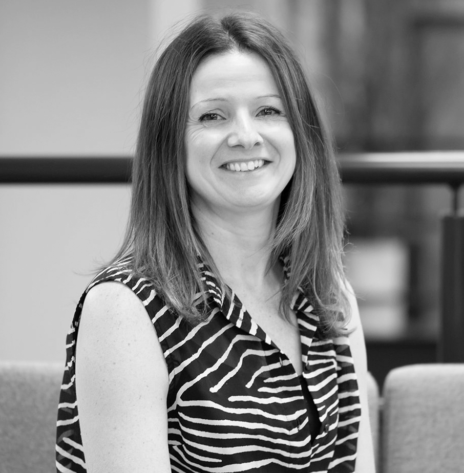 A black and white headshot image of a smiling woman on a sofa in front of an office backdrop.