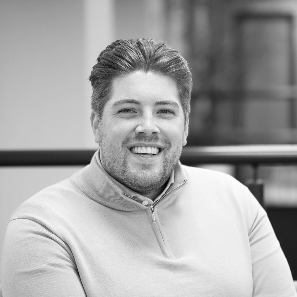 A black and white headshot image of a smiling man on a sofa in front of an office backdrop.