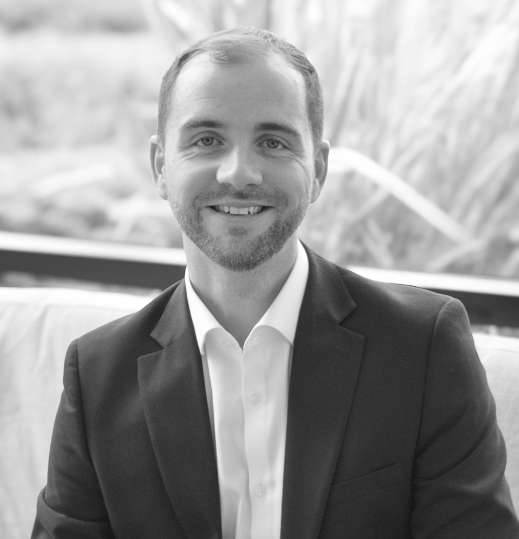 A black and white headshot image of a smiling man on a sofa in front of a window backdrop