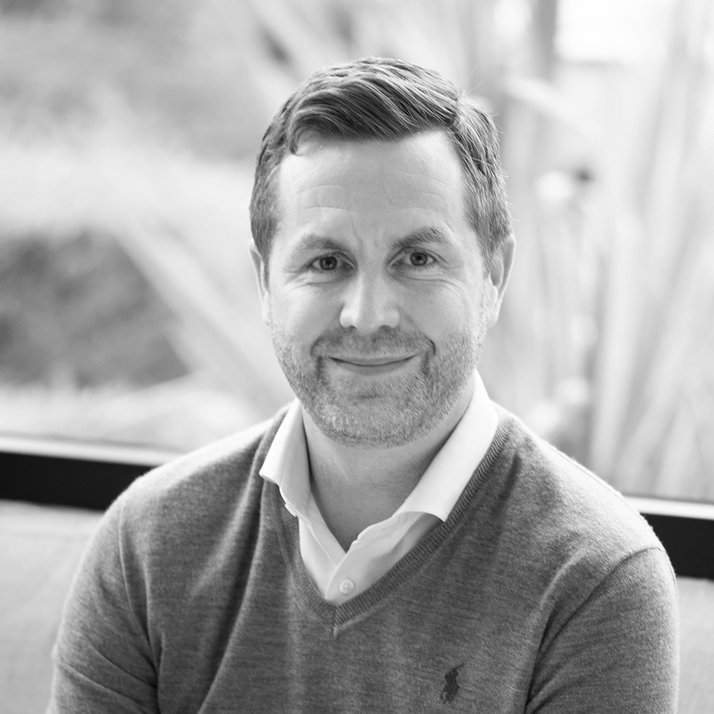 A black and white headshot image of a smiling man, Gregg Pendlington of Dow Schofield Watts, on a sofa in front of a window backdrop.