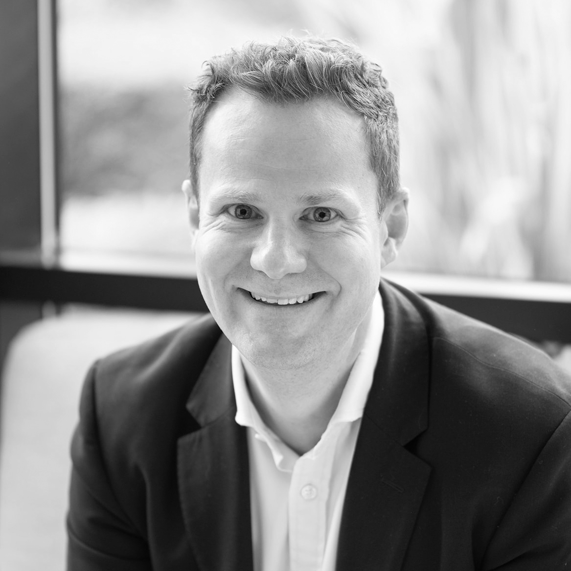A black and white headshot image of a smiling man on a sofa in front of a window backdrop.