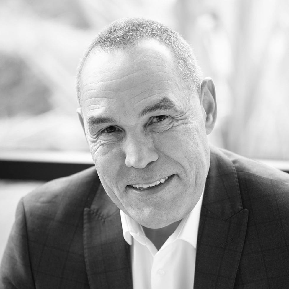 A black and white headshot image of a smiling man on a sofa in front of a window backdrop.