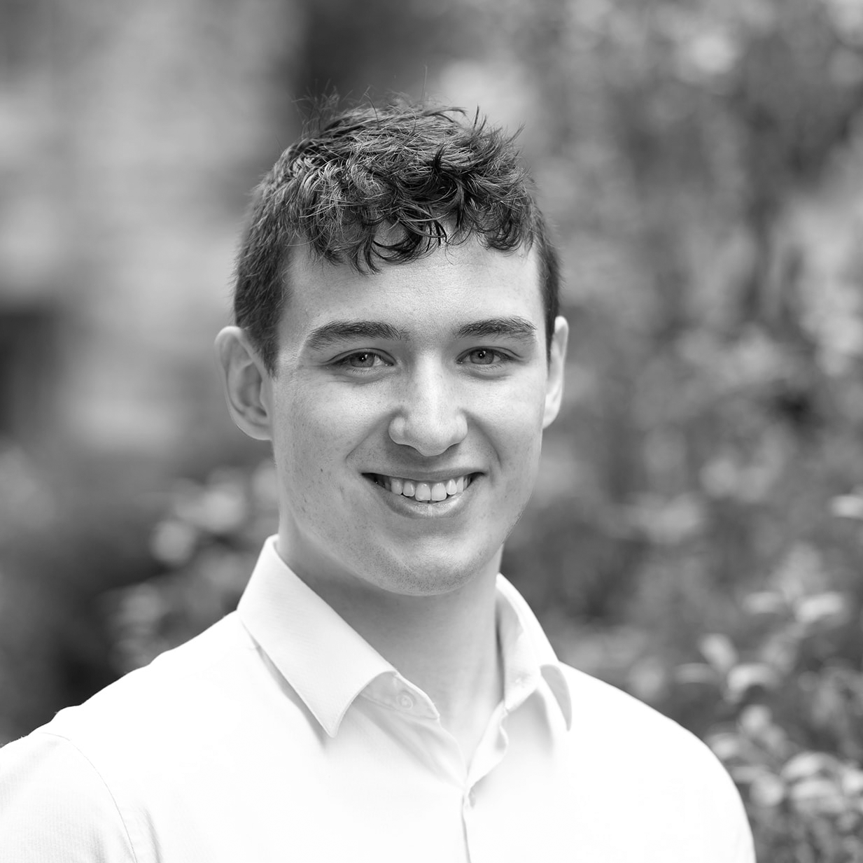 A black and white headshot image of a smiling man against a garden backdrop.