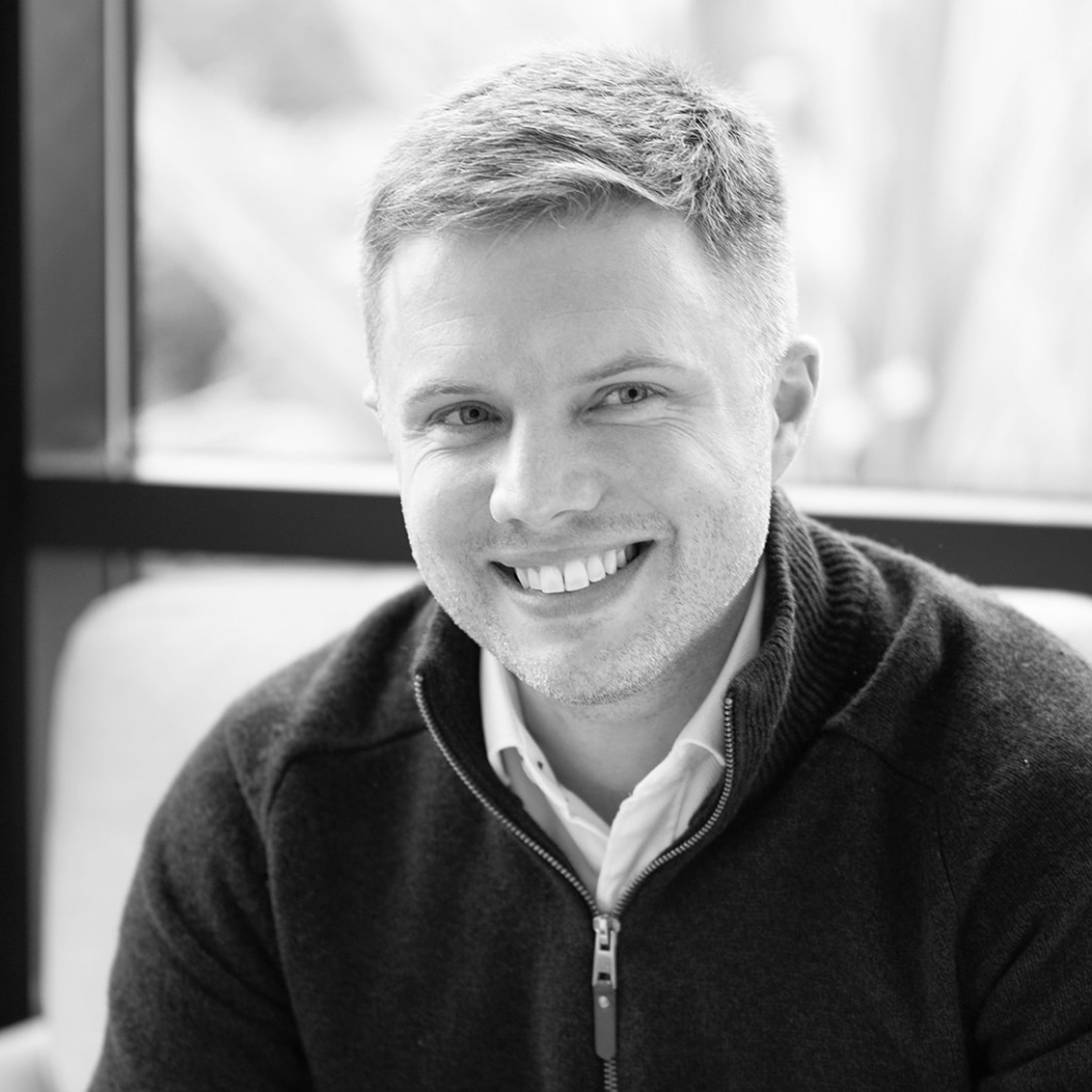 A black and white headshot image of a smiling man against a backdrop with a window.