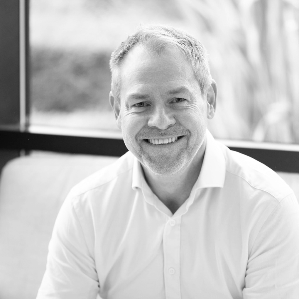 A black and white headshot image of a smiling man on a sofa in front of a window backdrop.