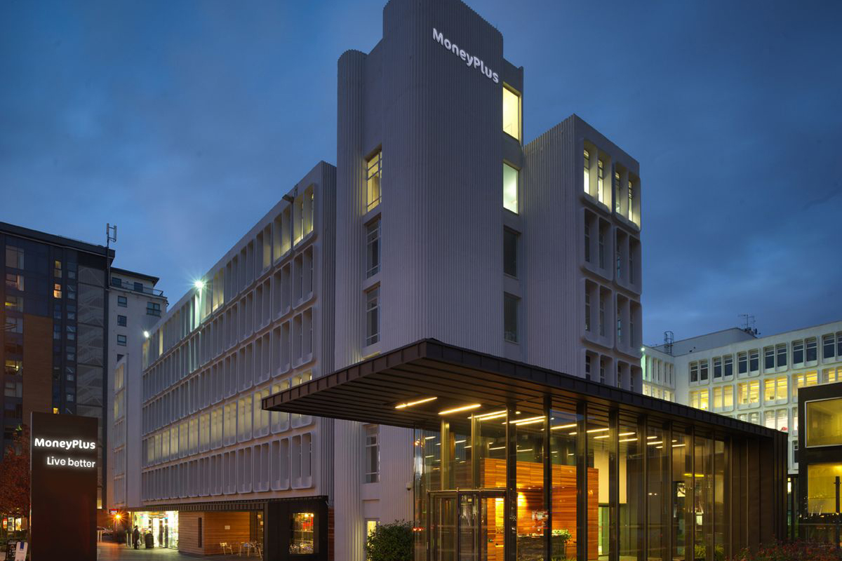 A modern multi-story office building with a "MoneyPlus" sign on top is shown at dusk.