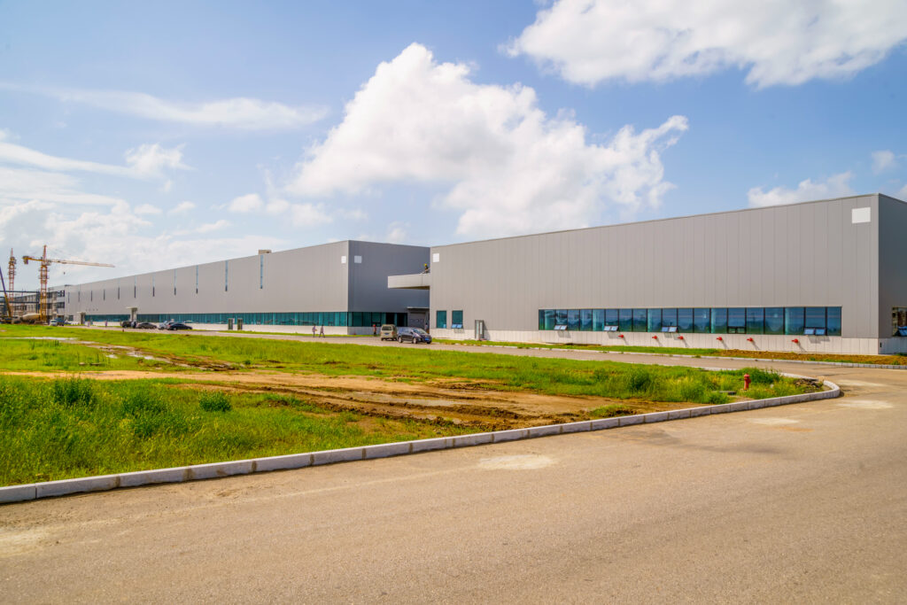 A large, modern, gray industrial warehouse or factory building sits under a bright blue sky with white clouds. A few vehicles are parked outside, and a patch of dirt and grass is in the foreground.