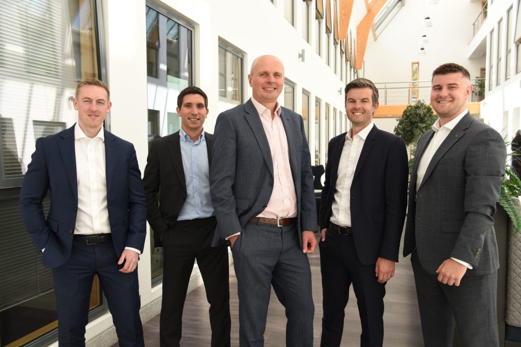 Five male co-workers smiling in an office setting, looking happy and comfortable.