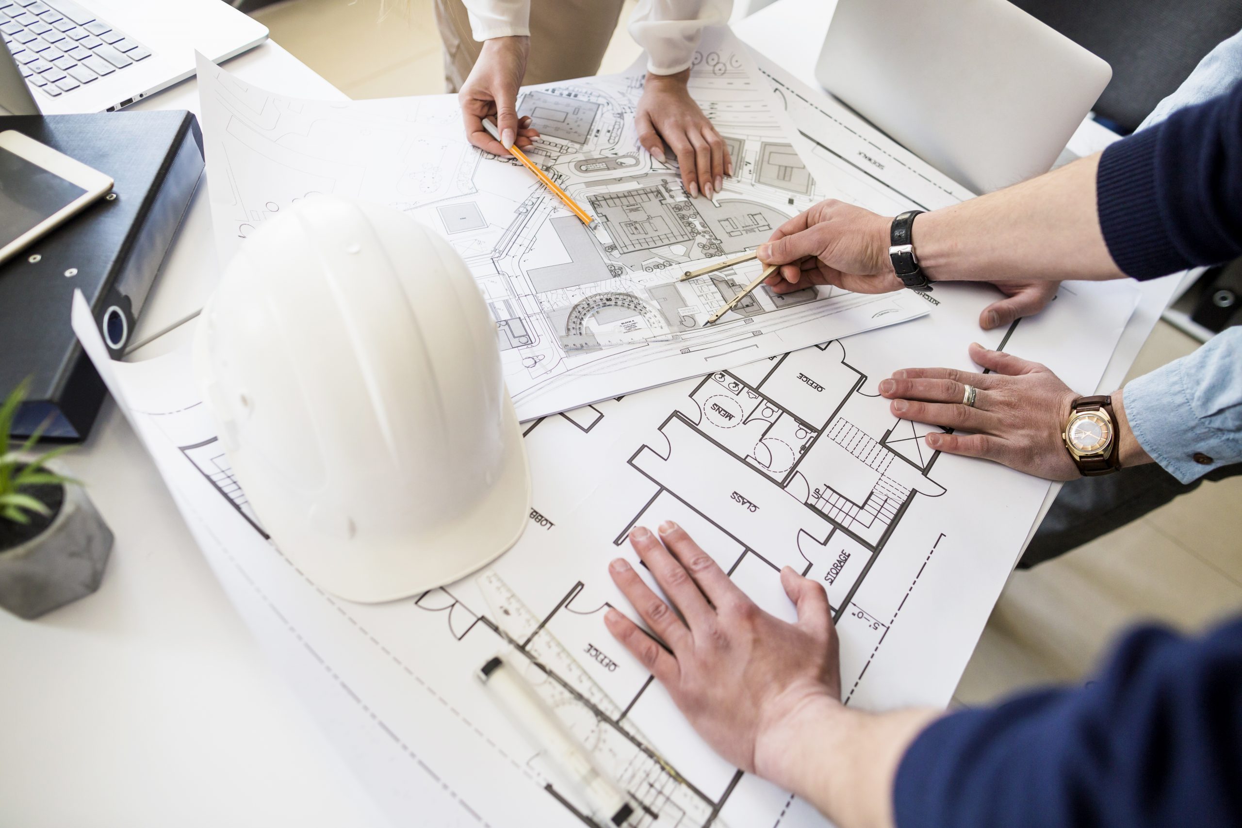 An overhead view of a group of architects or engineers collaborating on blueprints on a desk with a white hard hat.