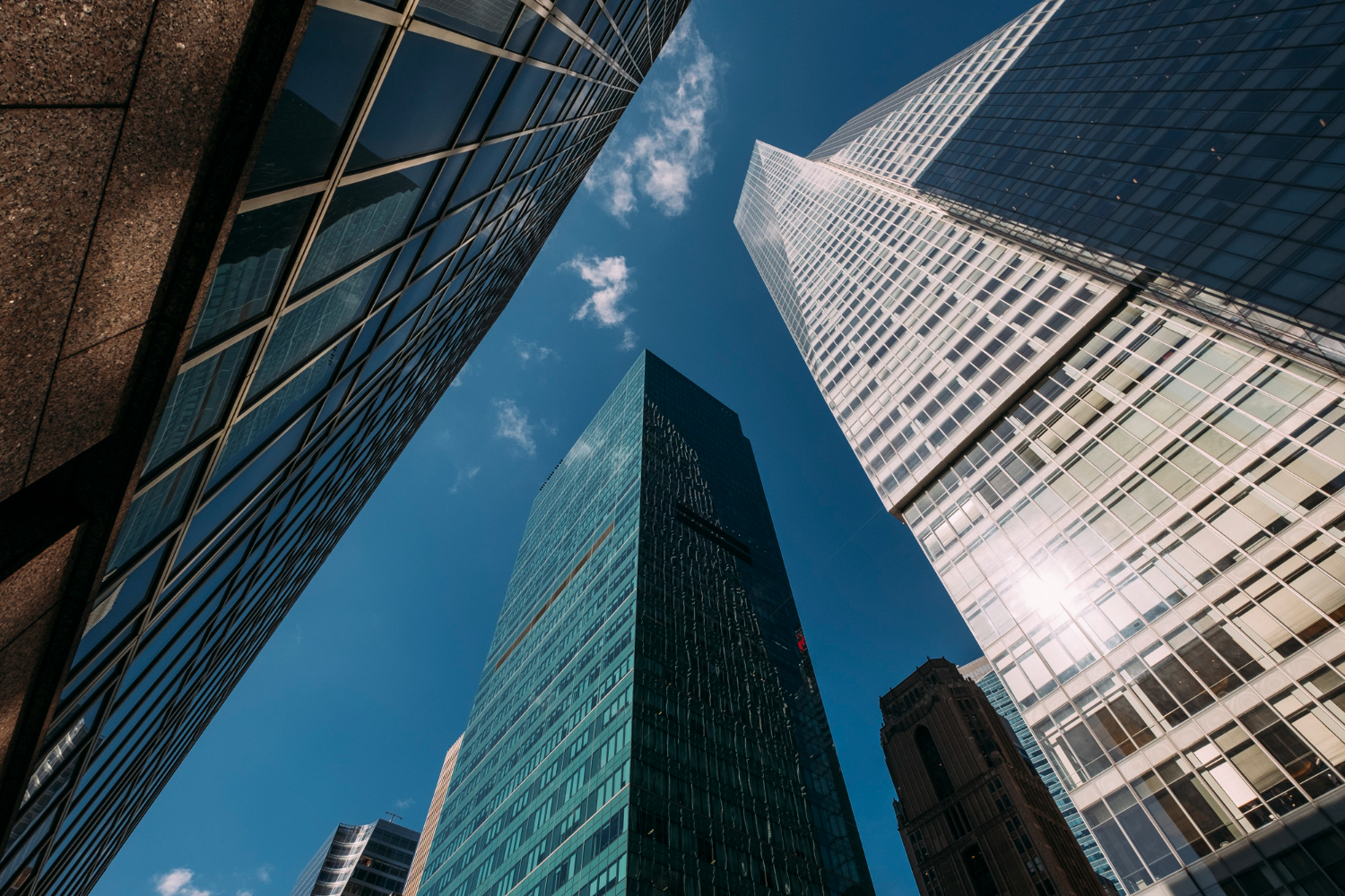 A low-angle view looking up at several modern skyscrapers with glass and metal facades, reflecting a bright blue sky with a few scattered clouds.
