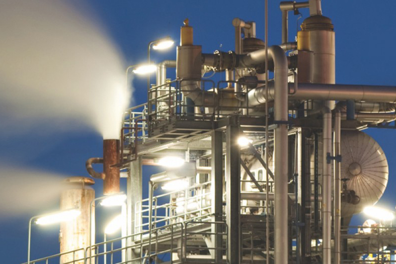A large industrial facility with pipes and towers is shown at night, with steam venting from a chimney.