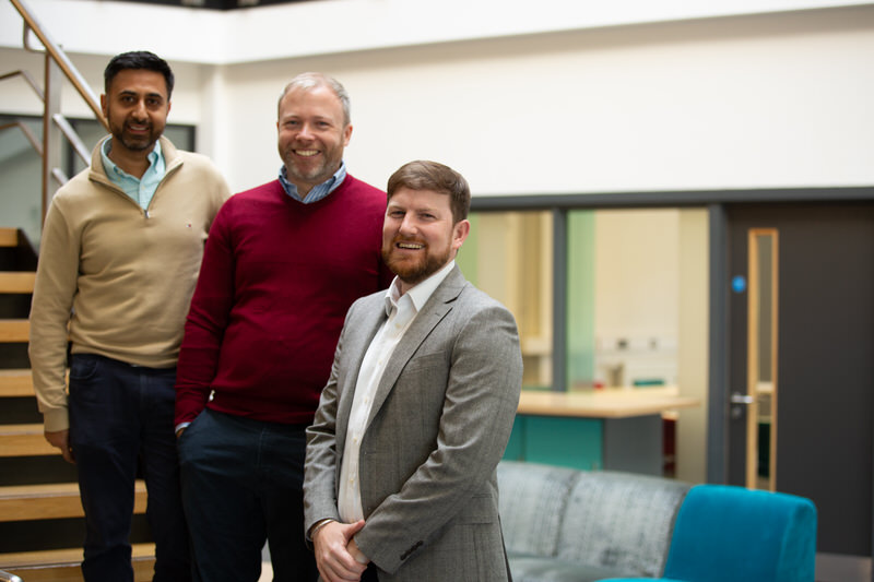 Three men stood smiling on a staircase in an office setting.
