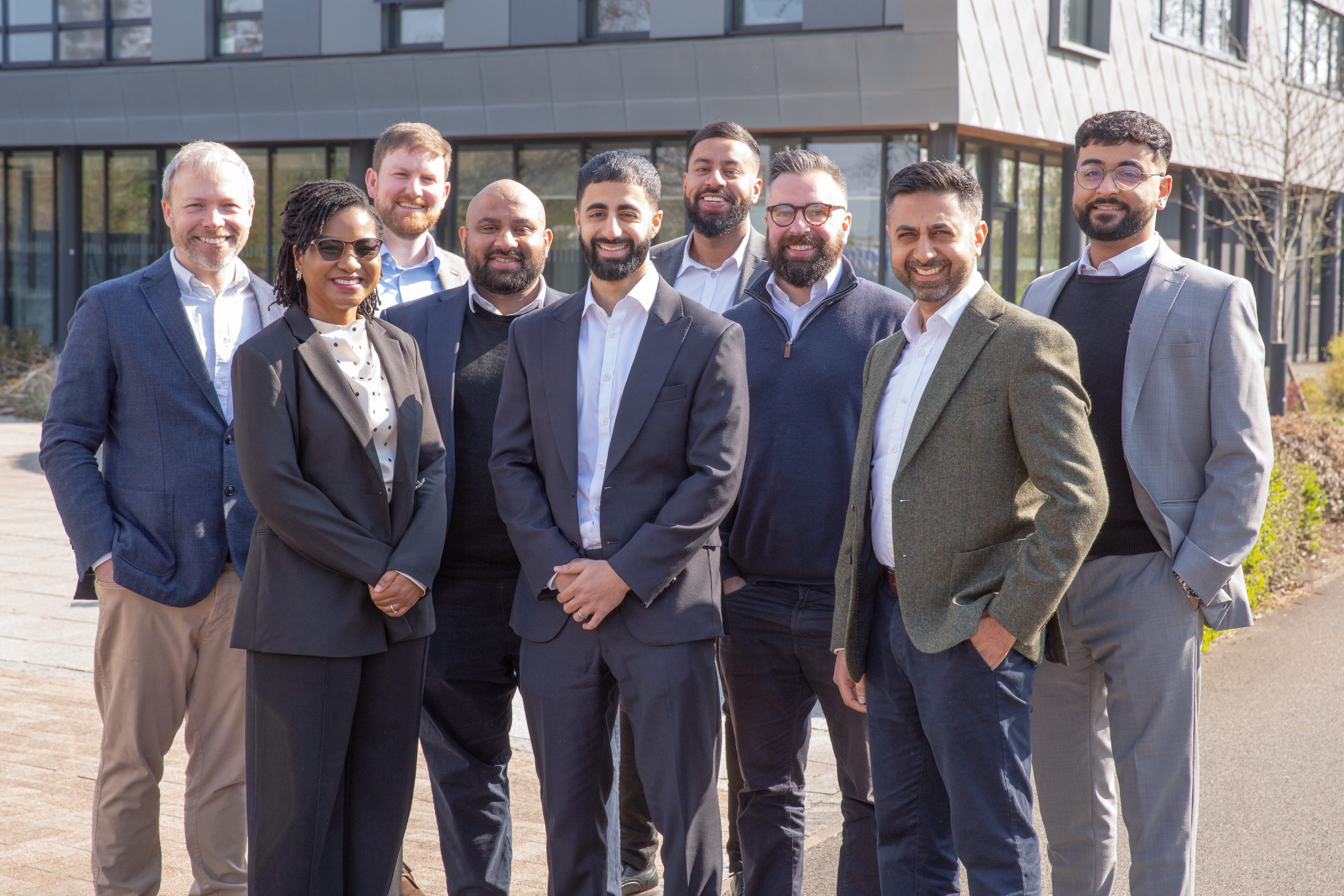 A group of nine smiling professionals of diverse ethnicities and genders are standing outdoors in front of a modern office building.