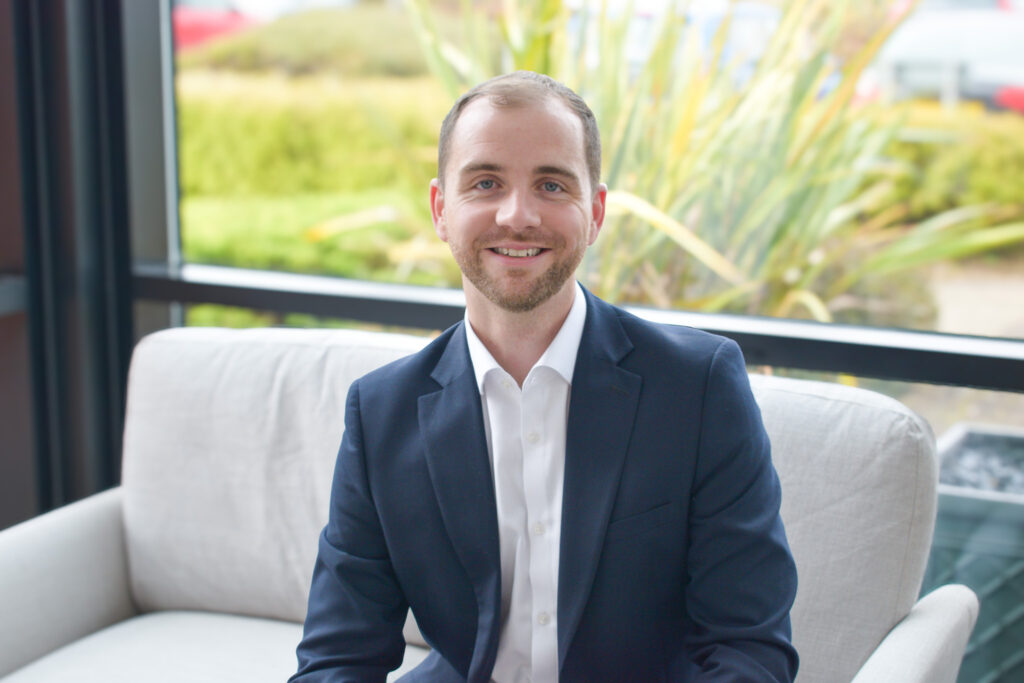 A headshot of a smiling Dan Walker on a sofa in front of a window backdrop.