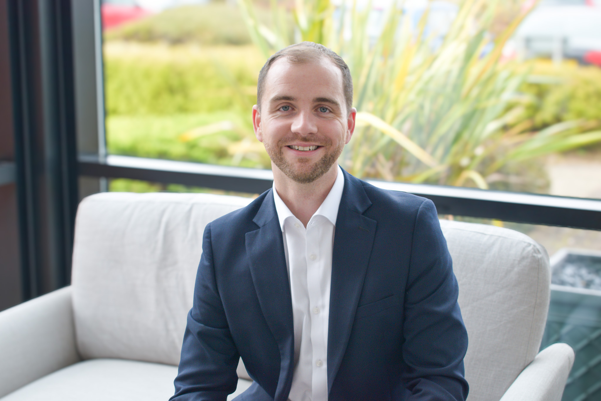 A headshot of a smiling Dan Walker on a sofa in front of a window backdrop.