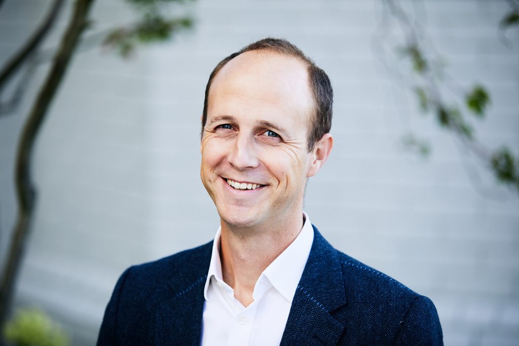 A headshot image of a smiling man against a garden background