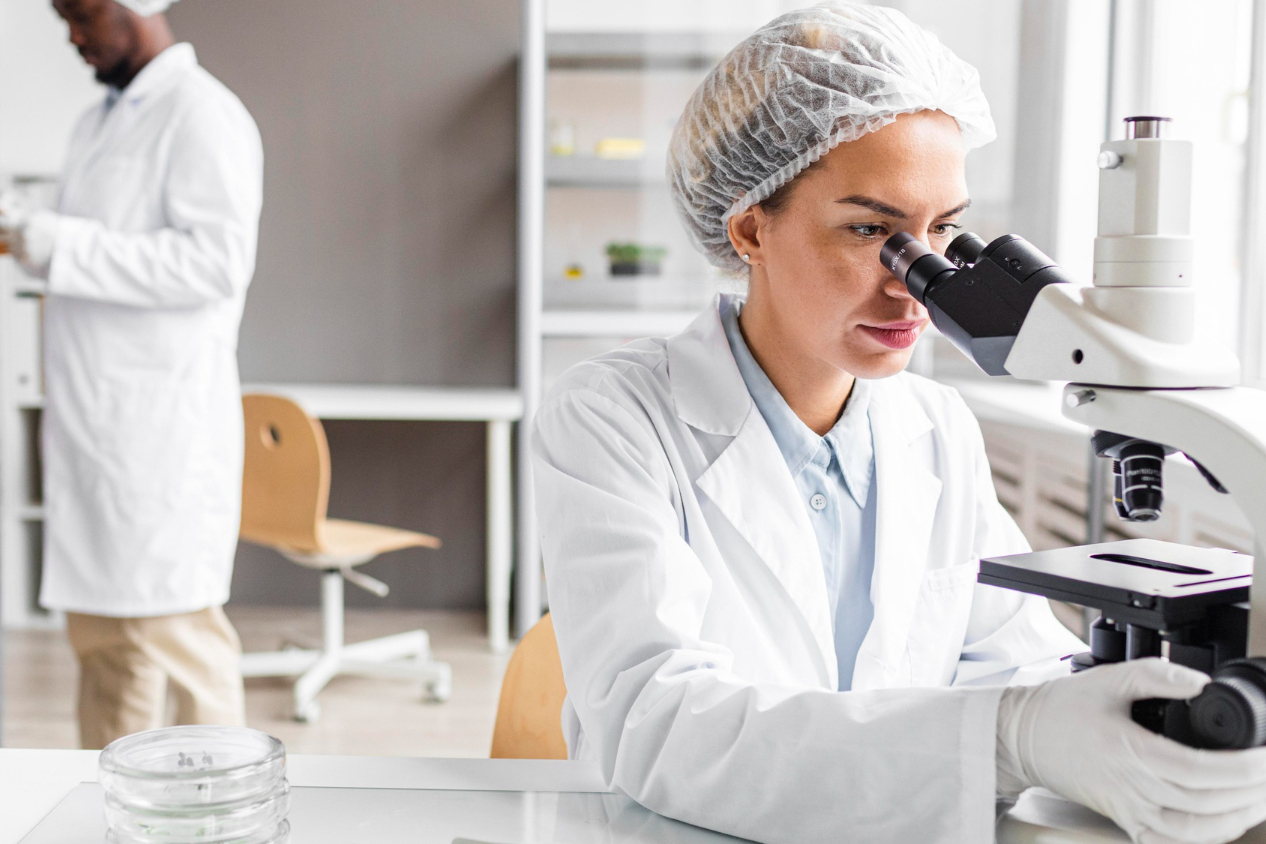 A female scientist wearing a lab coat and hair net looks through a microscope in a laboratory. Another scientist is in the background.