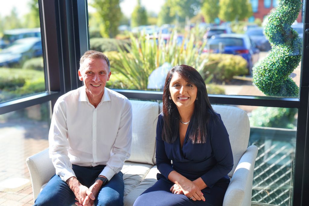 An image of a smiling woman and man on a sofa in front of a window backdrop