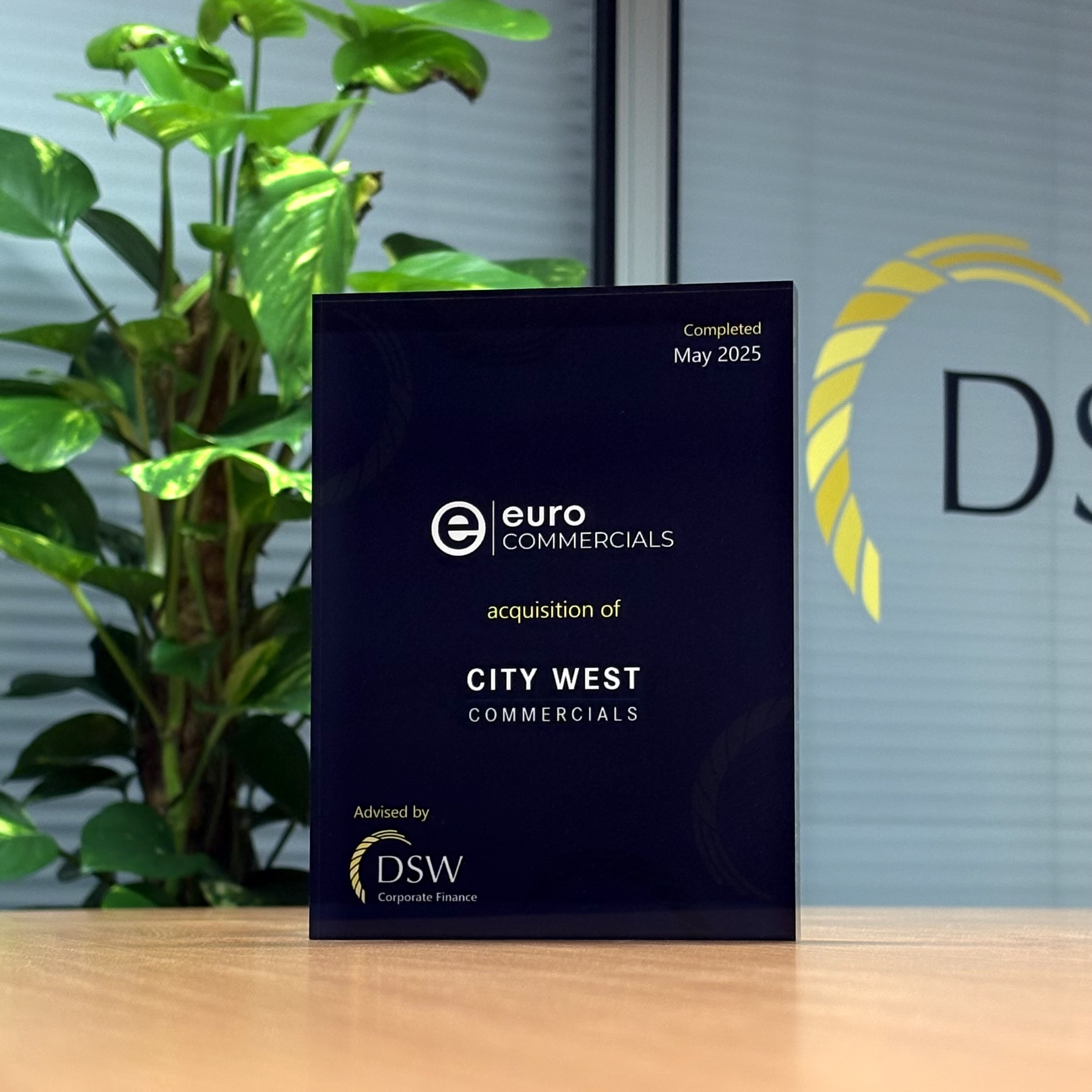 An AI-generated image of a plaque celebrating a corporate acquisition is sitting on a wooden desk. It features the logos for "Euro Commercials" and "DSW Corporate Finance" and commemorates the "acquisition of City West Commercials," completed in May 2025.