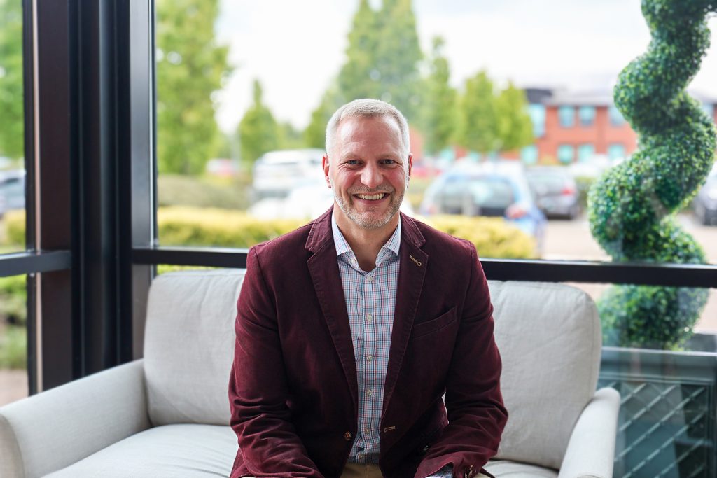 A smiling man, Paul Jesinger, on a sofa in front of a window backdrop.