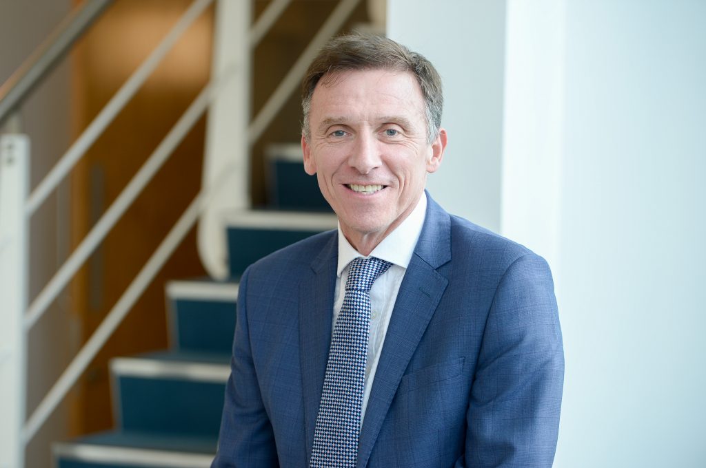 A headshot image of a smiling James Dow against an office staircase background