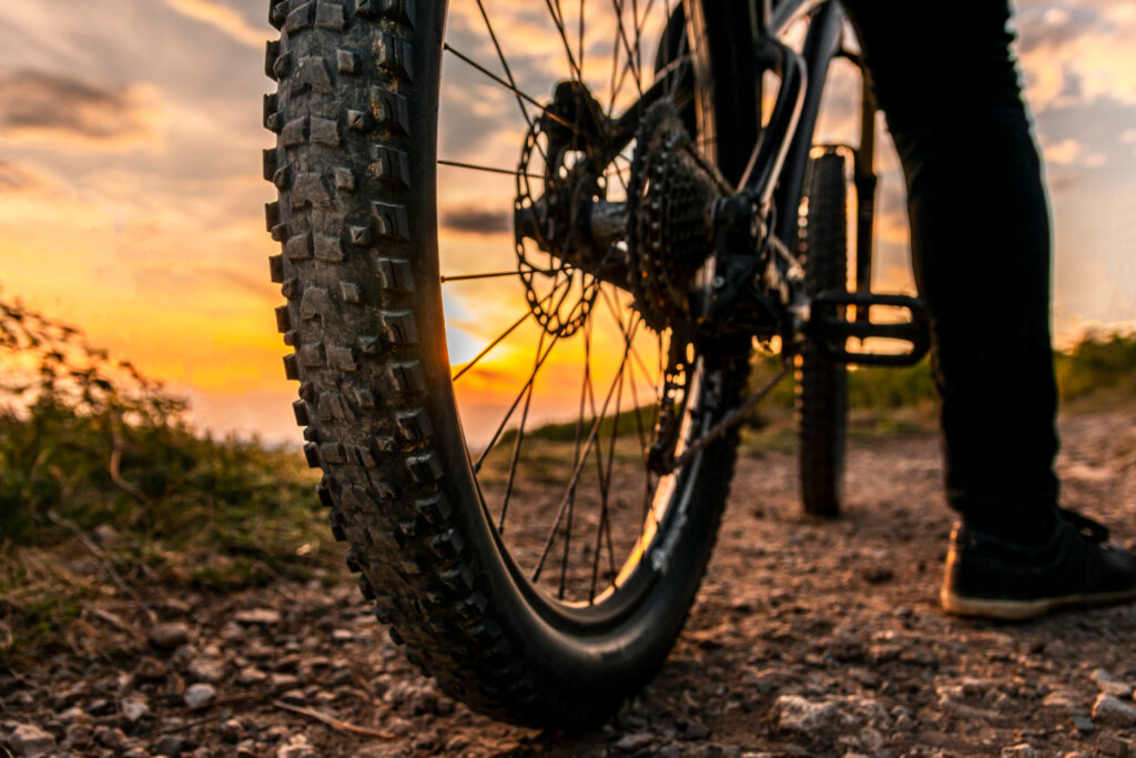 A close-up of a mountain bike wheel and pedals on a dirt trail at sunset.