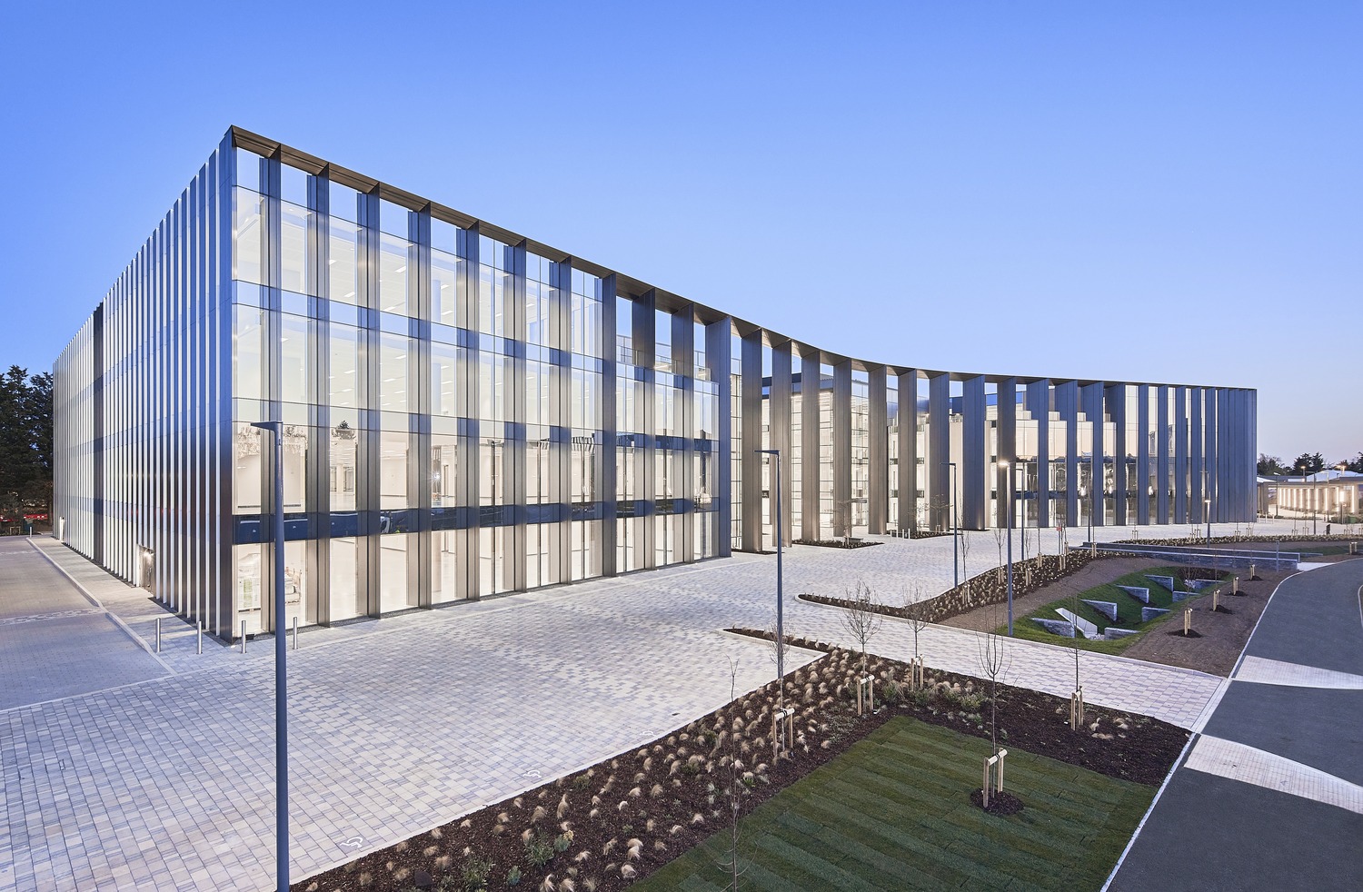 A low-angle exterior shot of a large, modern, glass-fronted building with a curved façade. The building has vertical fins for sun shading and is surrounded by a brick plaza and landscaped areas with small trees and grass.
