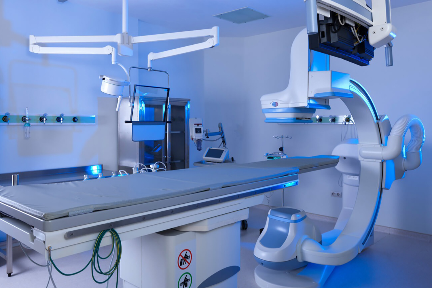 An empty operating room with a surgical table in the centre and a large, arched medical imaging machine on the right. The room is filled with medical equipment and has blue lighting.