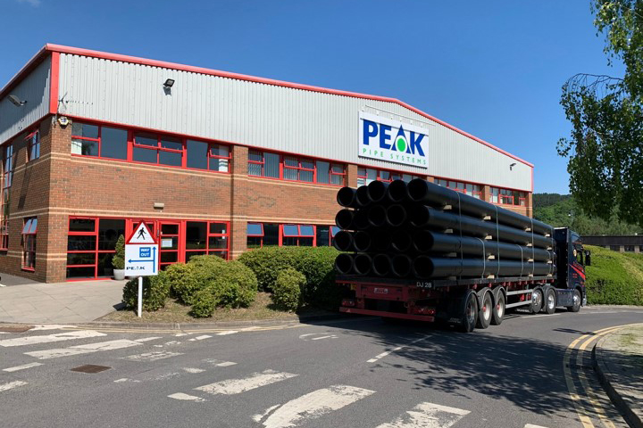 A truck with a flatbed trailer filled with large black pipes drives past the front of a red brick building with a sign that reads "Peak Pipe Systems."