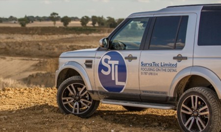 A silver SUV from "SurvaTec Limited" is parked on a dirt road at a dusty excavation or quarry site, with the logo visible on the side.
