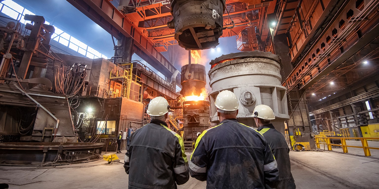 Three workers in hard hats stand in a large, industrial building with molten metal glowing in a vat.