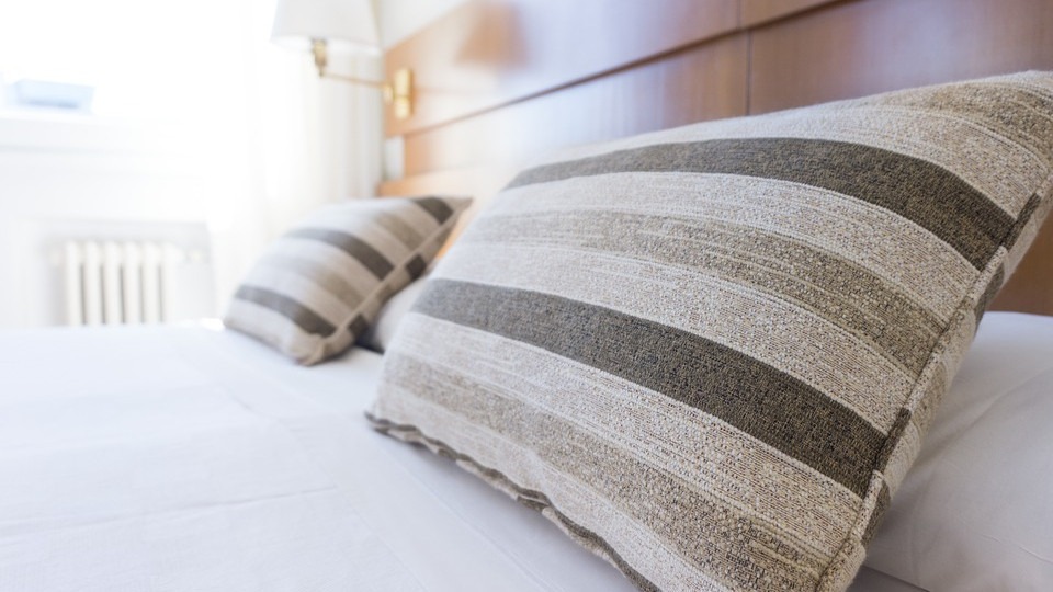 A close-up of a bed with white sheets and two striped pillows. The headboard is brown, and a radiator is visible in the background.
